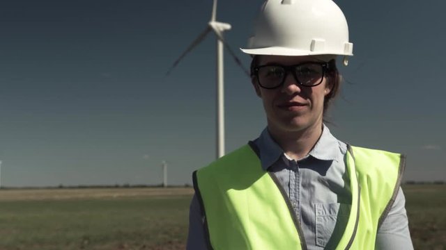 Portrait Of A Positive Successful Young Beautiful Woman Engineer In A Protective Helmet With Green Vest Standing Against A Background Of Windmills