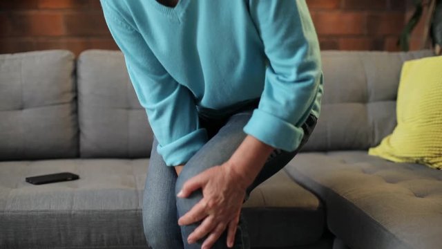 Attractive tall Caucasian old lady wearing casual clothes sitting on sofa. Cute grandma massaging leg. Pain in leg. Indoors. Room. Coziness.
