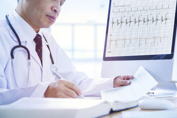 Asian doctor researches and analyses a patient’s health data on heartbeat graph in front of...