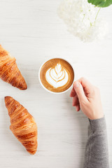 A cup of cappuccino with latte art and two croissants on white wooden table. Traditional french breakfast. Woman's hand holding a cup of coffee. Top view.