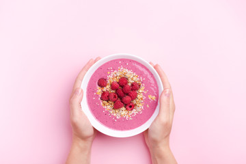 Woman's hands holding raspberries smoothie bowl on pink background. Top view, copy space.