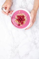 Woman's hands holding raspberries smoothie bowl on marble background. Top view, copy space.