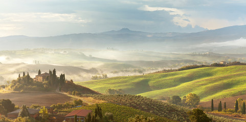 A beautiful Italian traditional rural landscape with autumn fields in the hills and a farmhouse with cypress and olive trees in a foggy morning at dawn - obrazy, fototapety, plakaty