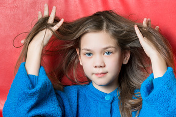 Cute girl in blue blouse tries to comb her hair with fingers on a red background