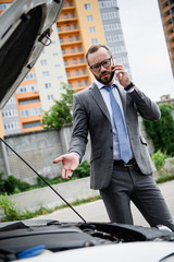 handsome businessman talking by smartphone near broken car with open hood