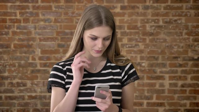Young sweet blonda is typing message on smartphone, dreaming concept, communication concept, brick background