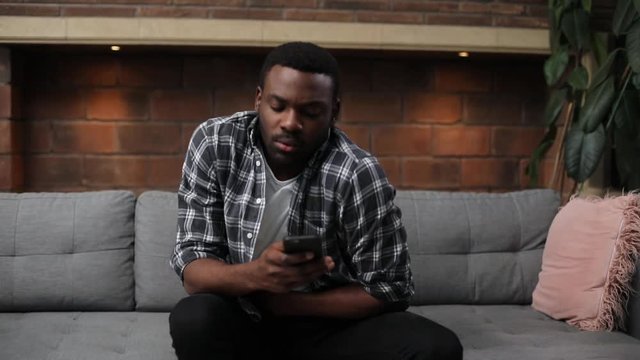 Cute African guy texting with girlfriend, sitting on sofa. Portrait of sick likable young man wearing plaid shirt. Stomachache. Indoors.