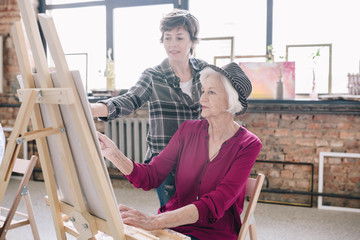 Portrait of elegant senior woman painting sitting at easel in art studio and enjoying art with smiling female teacher giving comments  against big windows.