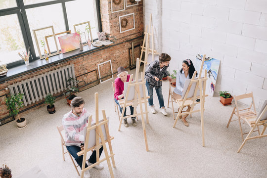 Above view portrait of students  painting sitting by easels in art studio with female art teacher watching them in spacious sunlit loft space, copy space