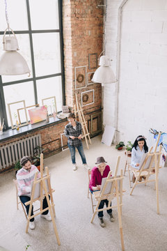 High angle view portrait of female art teacher working with group of students painting at easels in spacious sunlit art studio, copy space