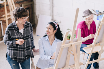 Portrait of pretty young woman painting sitting at easel in art studio with smiling female teacher giving comments, copy space