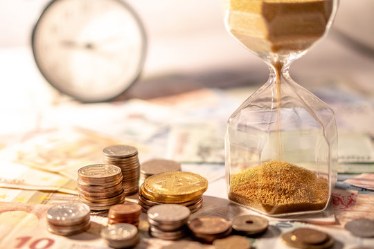 Sand running through the shape of hourglass on table with banknotes and coins of international currency. Time investment and retirement saving. Urgency countdown timer for business deadline concept