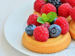 Soft sugar cookies topped with fresh forest fruits, raspberries, blackberries, blueberries on white plate, macro closeup