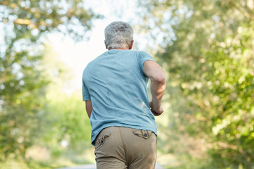 Naklejka na ściany i meble Back view of mature male joggs outside, has morning workout, enjoys sunny day and fresh air, being in motion, has healthy lifestyle. Elderly people, sport, motivation and retirement concept.