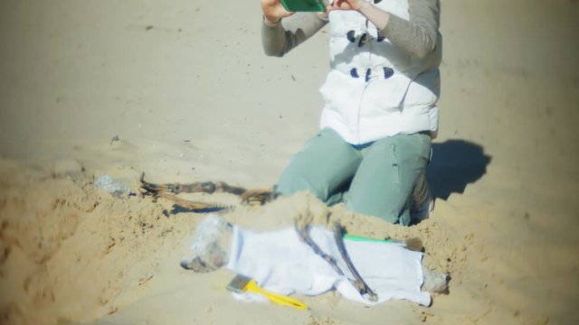 The woman is engaged in excavating bones in the sand, Skeleton and archaeological tools. Makes photo on smartphone