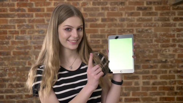 Young happy blonda is showing green screen of tablet, like sign, communication concept, brick background