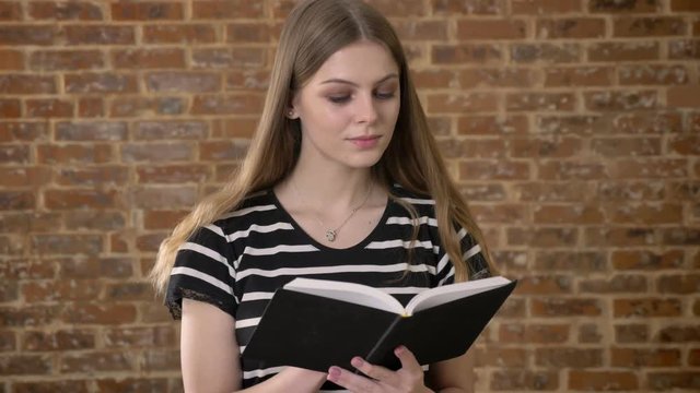 Young sweet blonda is reading book, watching at camera, smiling, brick background