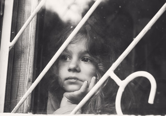 Black and white portrait of Sad little girl looks through window with lattice