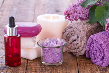 Red bottle with aromatic oil, soap, burning candle, bowl with sea salt, lilac flowers and towels