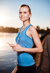 The girl in a t-shirt and shorts is standing by the lake with a smartphone and headphones after jogging.