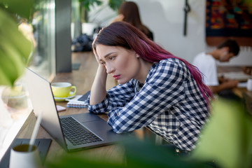 Fototapeta premium Side view of young businesswoman sitting at table in coffee shop. On table cup of coffee and laptop. In background white wall and window.