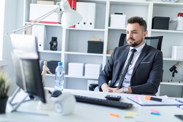 A man sits in a chair in the office at the table and looks at the monitor.
