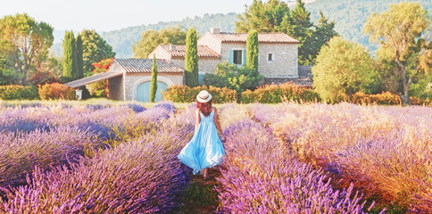 Schönes Mädchen, das sich in blauem Boho-Chic-Kleid und Strohhut anzieht, das ein erstaunliches blühendes Lavendelfeld in der Provence, Frankreich, geht. Panoramablick. Postproduktionsfoto in traditionellen provenzalischen Pastelltönen.
