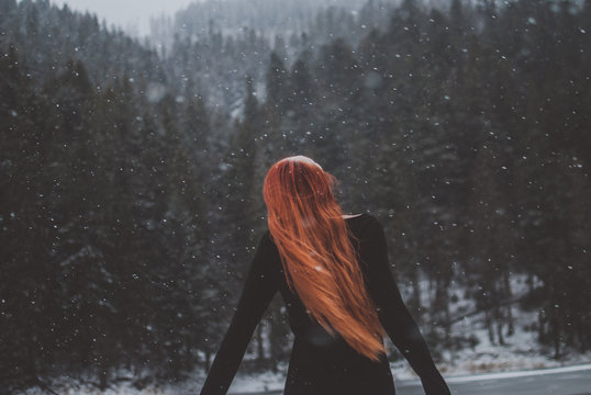 Handsome dark-eyed redhead girl pretending she's flying by mountain lake called Synevyr and looking up in the distance. Snowflakes descending on her beautiful hair. 