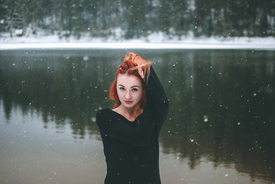 Snowflakes flying over handsome black-eyed redhead girl looking at the camera and playing with her beautiful hair. Closeup of falling snowflakes. Splendid shady black mountain lake reflecting trees