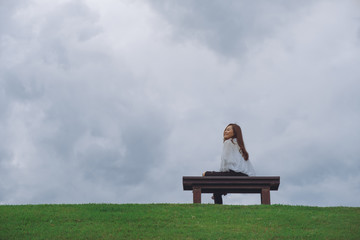 A happy woman sitting on a wooden bench in the park with cloudy and gloomy sky background