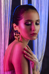 A beautiful glamorous brunette girl with naked shoulders wearing large elegant earrings peeps through a translucent curtain made of white threads. Multicolored artistic stage lighting.