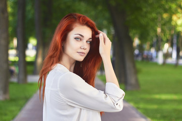 Fototapeta premium Close up of attractive red-haired girl in white shirt with long foxy hair posing in park background. Ginger teenager holds her hand near the head looking at camera.