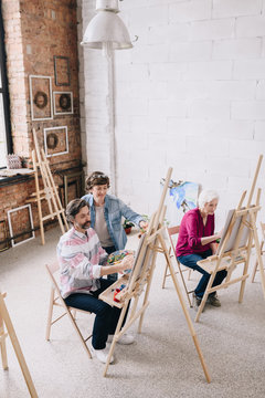 High angle portrait of adult students  painting sitting by easels in art studio with female art teacher , copy space