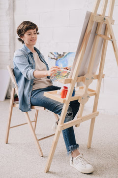 Full length portrait of smiling female artist painting sitting by easel  in art studio