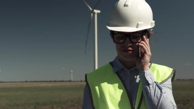 Portrait Of An Attractive Young Woman Engineer In Glasses In A Protective Helmet And Green Vest Talking On A Smartphone With The Project's Curator Against The Background Of Windmills