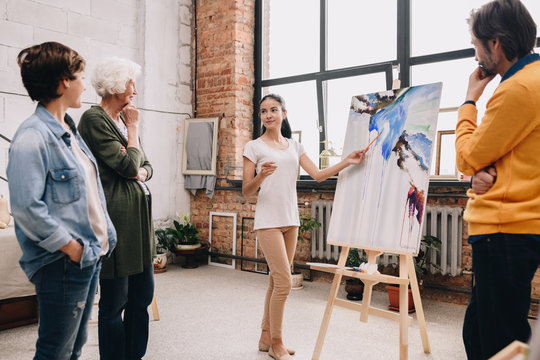 Full length portrait of pretty young woman presenting modern watercolor painting to audience standing in art-studio or gallery