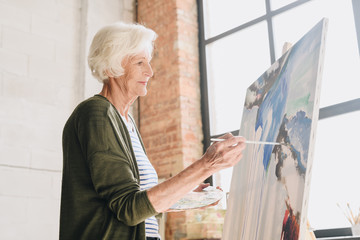 Side view portrait of white haired senior woman holding palette painting pictures at easel in  art...