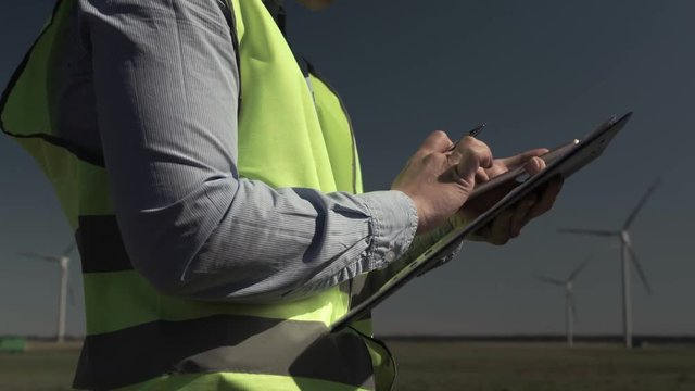 Hands Of An Unidentified Engineer In The Green Vest Are Holding A Smartphone And A Folder With A Work Plan On The Background Of Windmills. Concept Of Alternative Energy