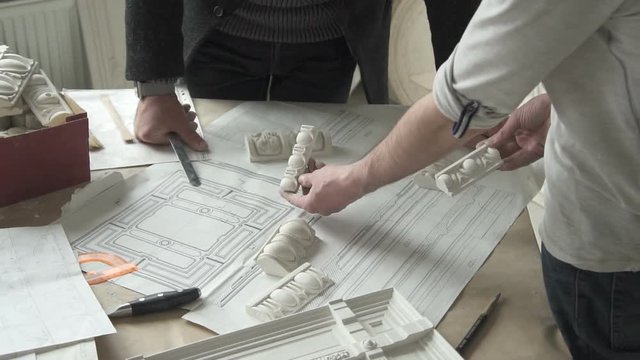 Hands of male sculprot and architector comparing white plaster molding samples with paper draft on the table in bright studio. Indoors.