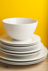 Big stack of different white plates, bowls, isoalted yellow background, one above the another. Wooden table, Vertical Image Copy Space.