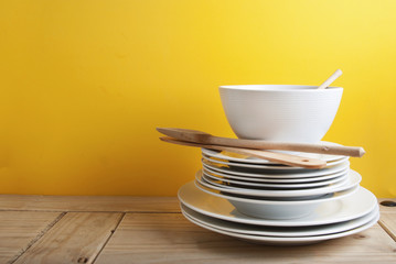 Big stack of different white plates, bowls, isoalted yellow background, one above the another. Wooden table, Copy Space.