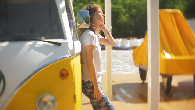 young woman outdoors by yellow vagon car listening music in headphones using smartphone - relaxing, enjoying, concept of technology and travel