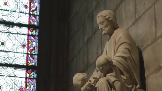 Handheld moving shot of Jesus' statue with children and stained windows in background inside an old french church