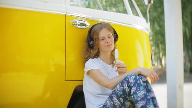 young woman outdoors by yellow vagon car listening music in headphones using smartphone - relaxing, enjoying, concept of technology and travel