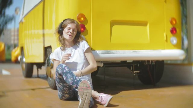 young woman outdoors by yellow vagon car listening music in headphones using smartphone - relaxing, enjoying, concept of technology and travel
