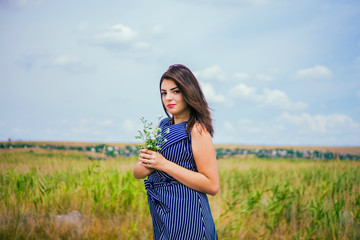 A brunette girl in a long blue dress with a bouquet of wildflowers in a summer field