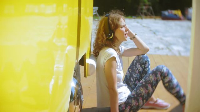 young woman outdoors by yellow vagon car listening music in headphones using smartphone - relaxing, enjoying, concept of technology and travel