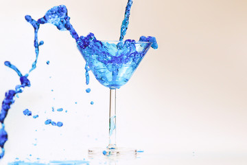 Cocktail with blue liquid in glass. Glass with blue water pouring with liquid with splashes and drops. Martini glass filling with alcohol with splashes on white background. Refreshing drink concept.
