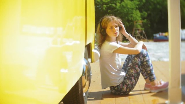 young woman outdoors by yellow vagon car listening music in headphones using smartphone - relaxing, enjoying, concept of technology and travel
