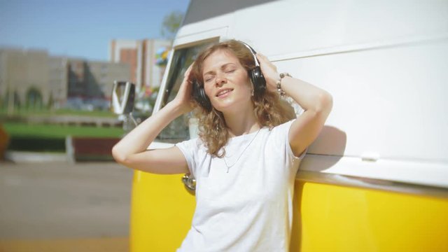 young woman outdoors by yellow vagon car listening music in headphones using smartphone - relaxing, enjoying, concept of technology and travel